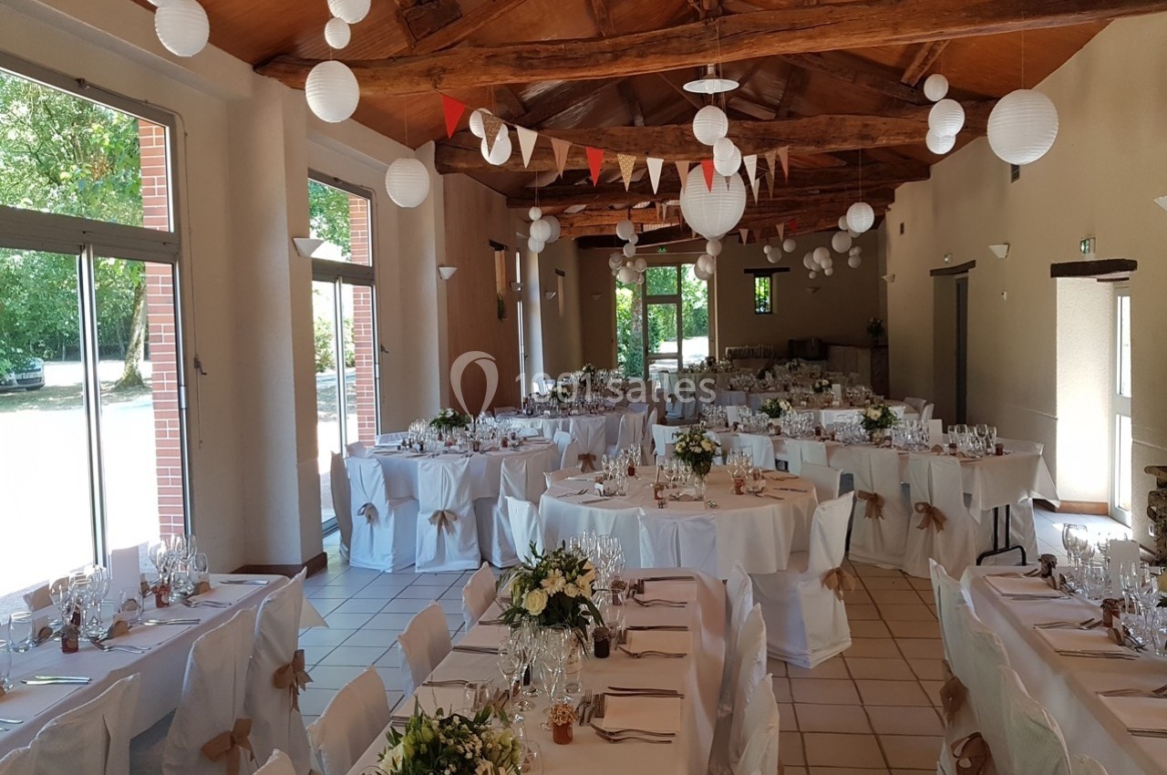 Salle de réception décorée pour un mariage, avec tables dressées, guirlandes et lanternes suspendues au plafond en bois.
