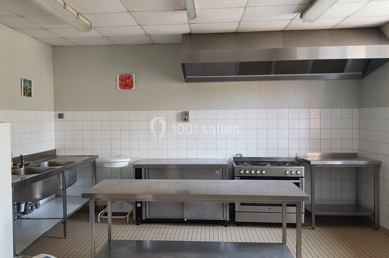 Cuisine professionnelle avec carrelage blanc, plan de travail en inox, évier, cuisinière et hotte.
