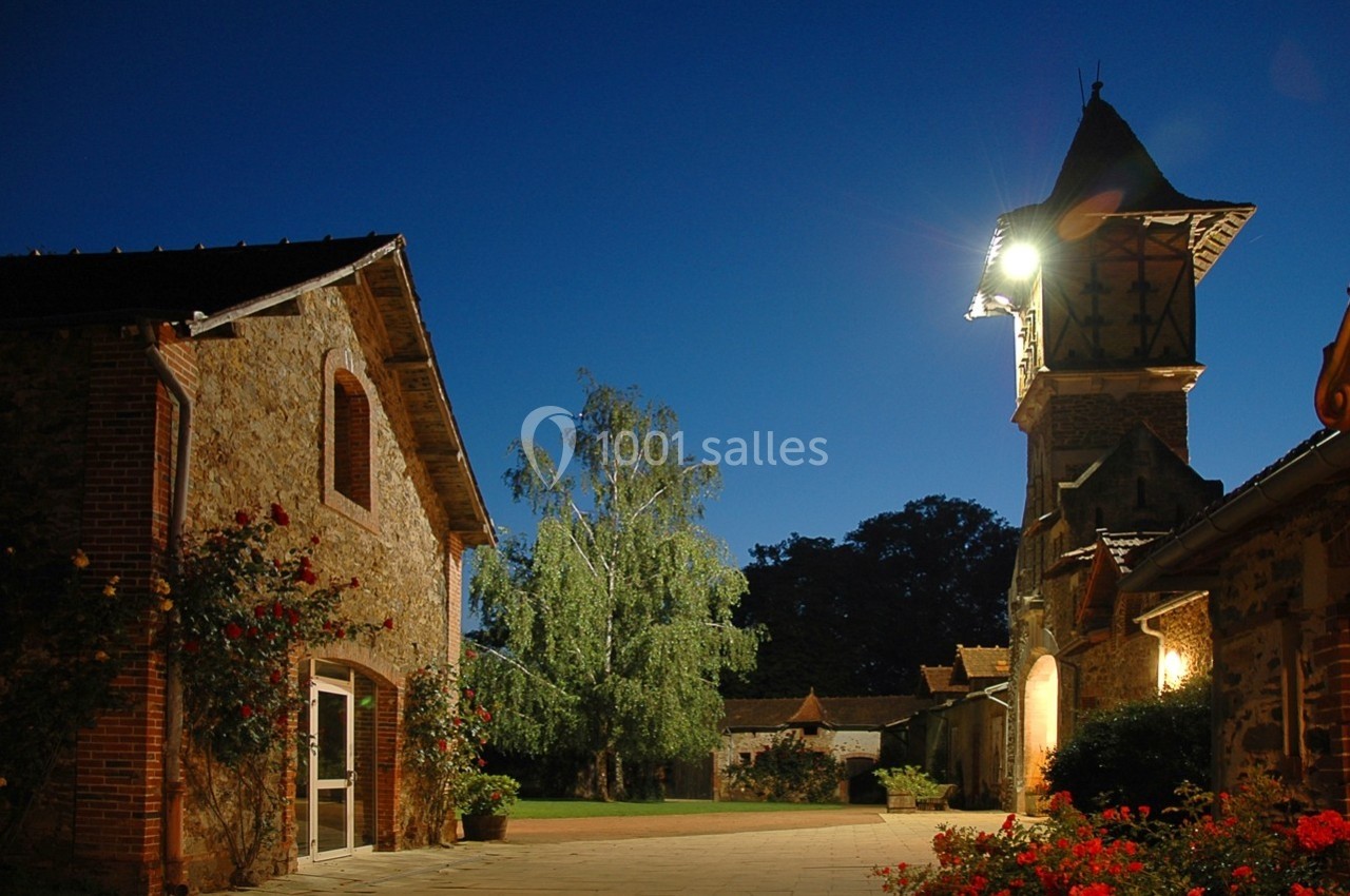 Cour pavée au crépuscule avec bâtiments en pierre, clocher éclairé et arbre au centre.