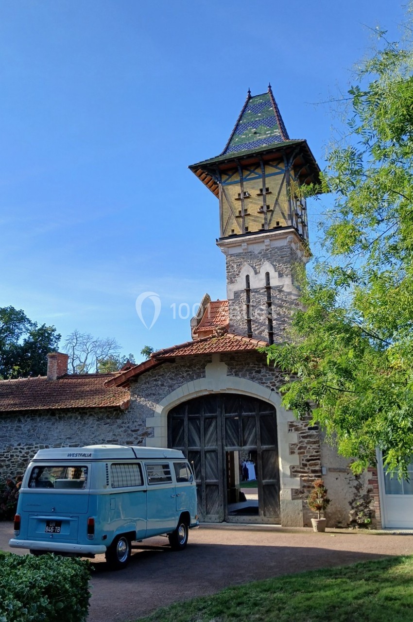 Un van bleu garé devant un bâtiment ancien avec une tour en pierre et un toit coloré, entouré de verdure.