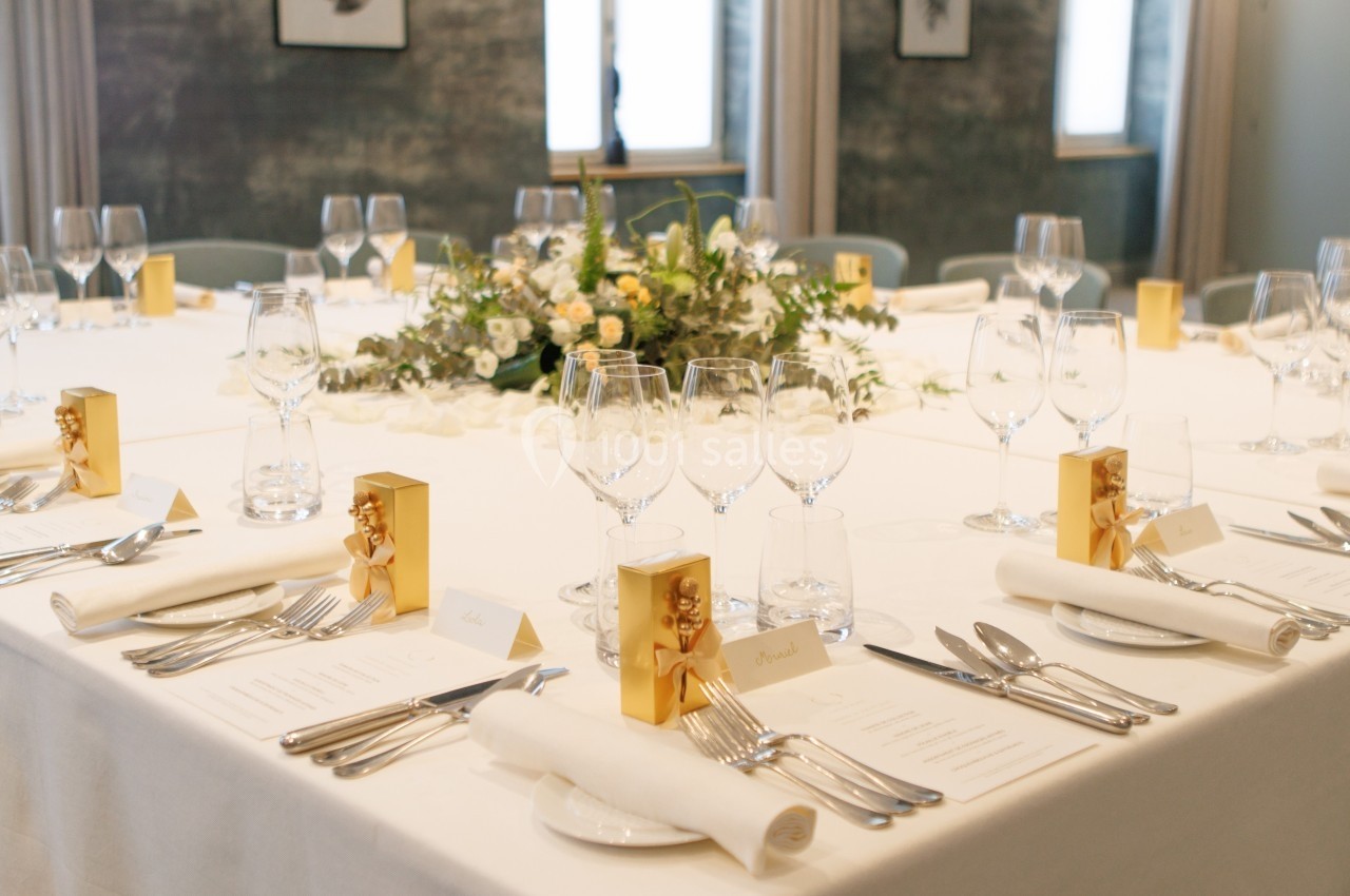 Table élégamment dressée avec nappes blanches, couverts, verres, menus, décor floral central et petits cadeaux dorés.