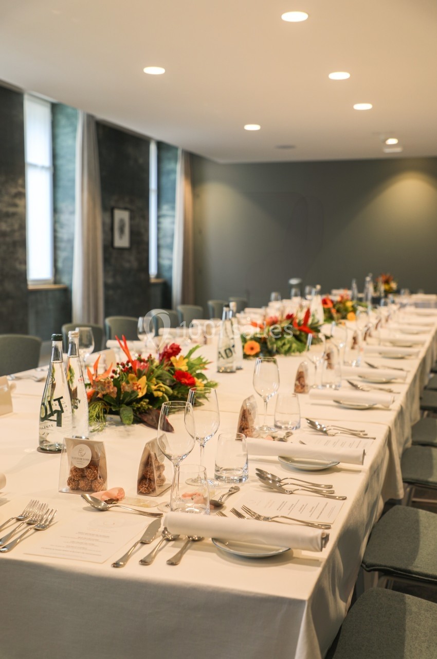 Table dressée pour un repas ou une réunion, avec nappes blanches, couverts, verres, menus et décorations florales.