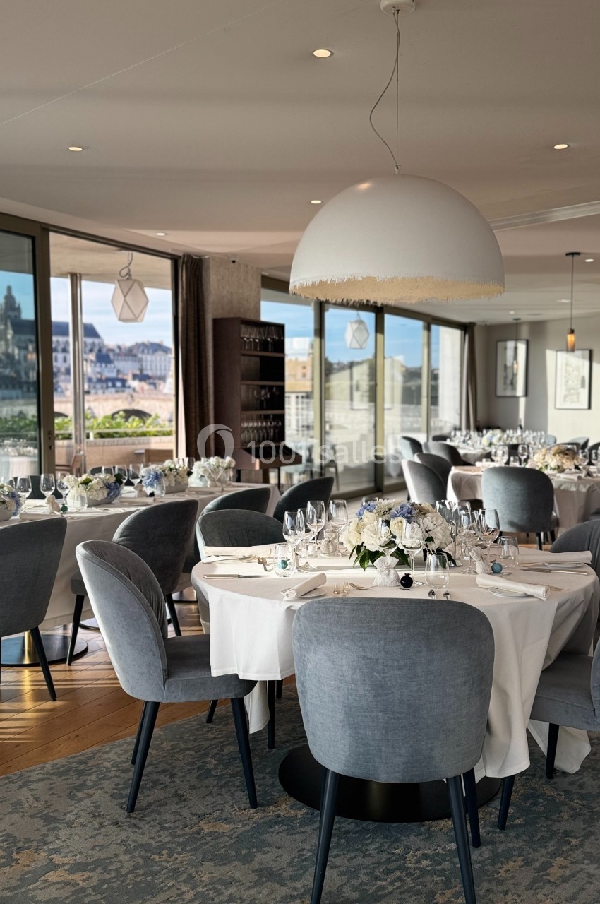 Salle de restaurant élégante avec tables dressées, chaises grises et vue panoramique sur une ville en arrière-plan.