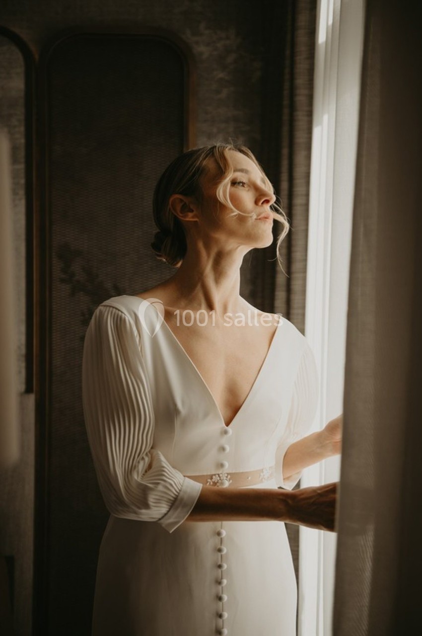 Femme en robe blanche élégante regardant par une fenêtre, éclairée par une lumière douce.