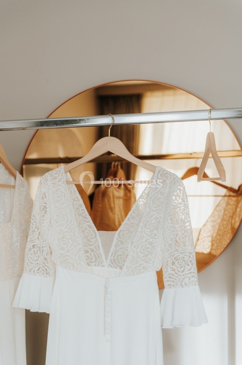 Robe de mariée blanche en dentelle suspendue sur un cintre devant un miroir rond doré.