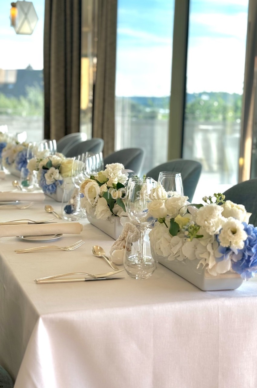 Table élégamment dressée avec nappes blanches, vaisselle raffinée et compositions florales bleues et blanches.