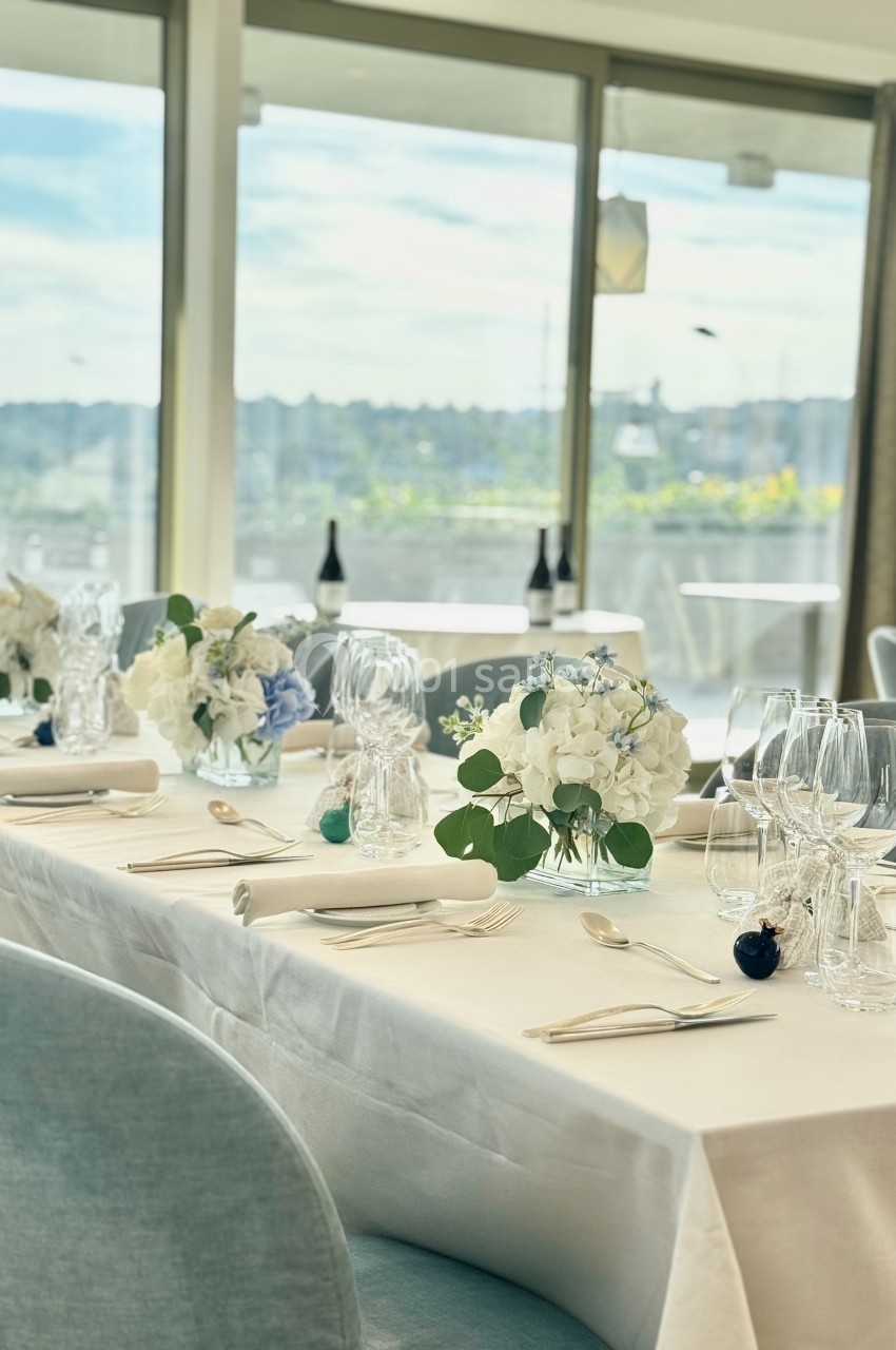 Table élégamment dressée avec nappes blanches, verres, couverts et bouquets de fleurs, devant une baie vitrée lumineuse.