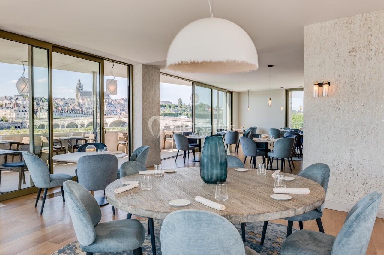 Salle de restaurant lumineuse avec grandes baies vitrées offrant une vue sur un pont et une ville en arrière-plan.