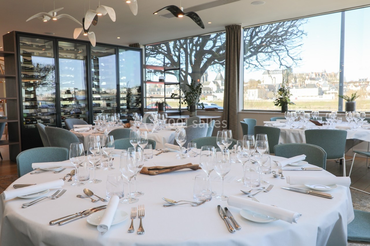 Salle de restaurant lumineuse avec tables élégamment dressées, grandes baies vitrées et vue sur un paysage urbain.