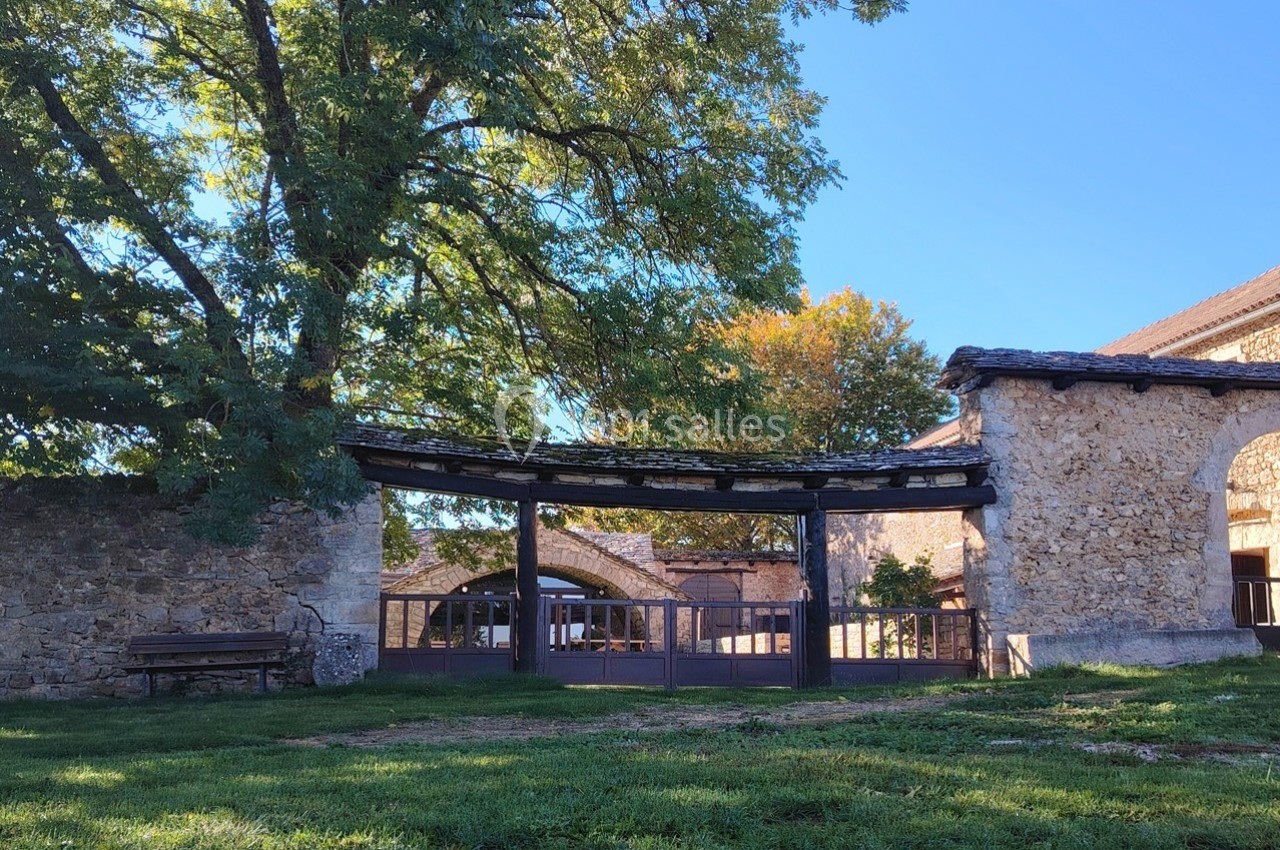 Entrée d'un bâtiment en pierre avec une arche en bois, entourée d'arbres et d'une pelouse verte.