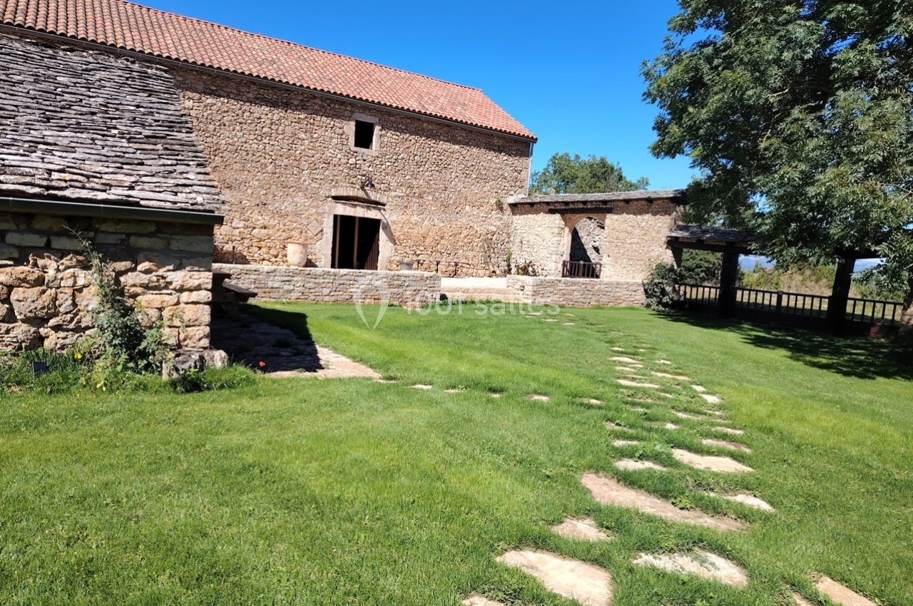 Cour d'une ferme en pierre avec bâtiments anciens, pelouse verte, chemin en dalles et arbre ombrageant.