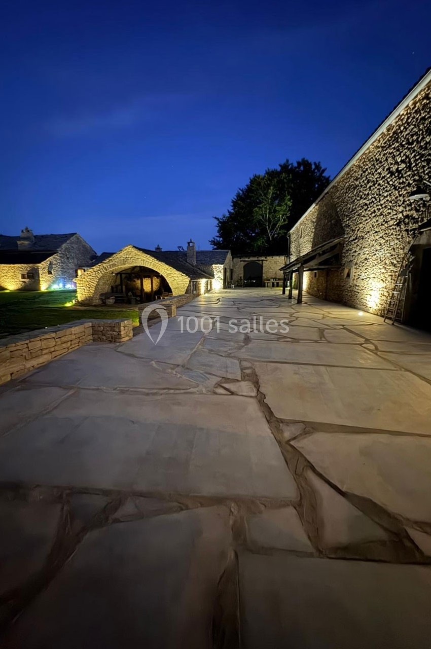 Cour en pierre éclairée la nuit, entourée de bâtiments en pierre avec des arbres en arrière-plan.