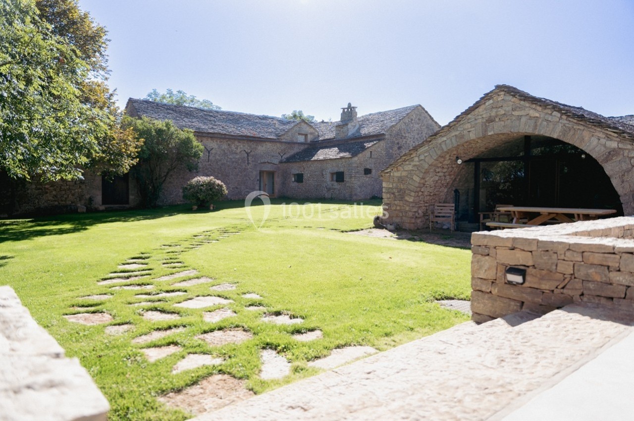 Cour verdoyante avec chemin en pierres menant à une bâtisse en pierre et une structure voûtée sous un ciel dégagé.