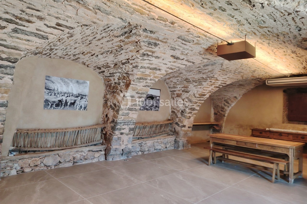 Salle voûtée en pierre avec bancs en bois, table en bois massif et éclairage doux au plafond.