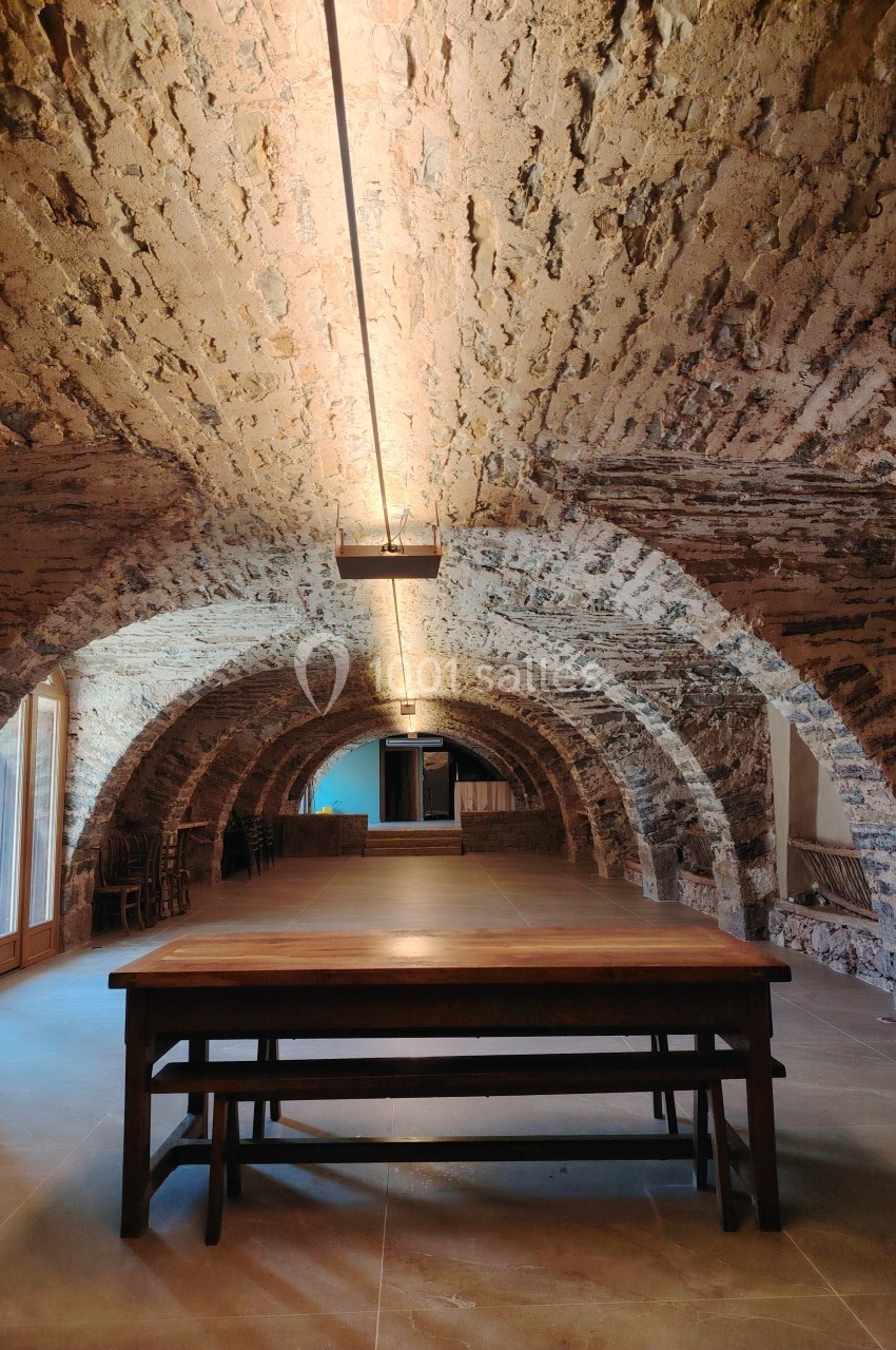 Salle voûtée en pierre avec une grande table en bois au centre, éclairée par des lumières suspendues.