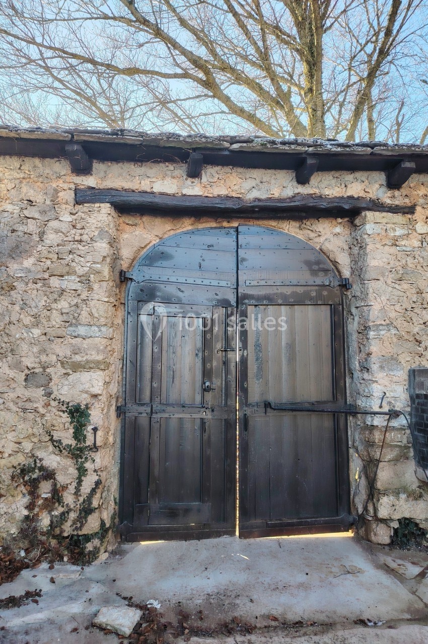 Porte en bois sombre avec des ferrures métalliques, encadrée par un mur en pierre ancienne sous des arbres dénudés.