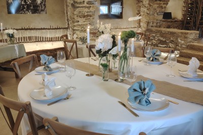 Salle voûtée en pierre avec une table ronde dressée, ornée de fleurs blanches et serviettes pliées bleues.