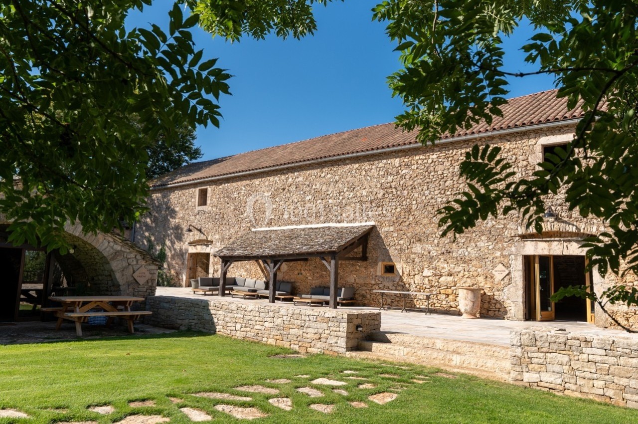 Bâtisse en pierre avec terrasse couverte, entourée de pelouse et d'arbres sous un ciel bleu.