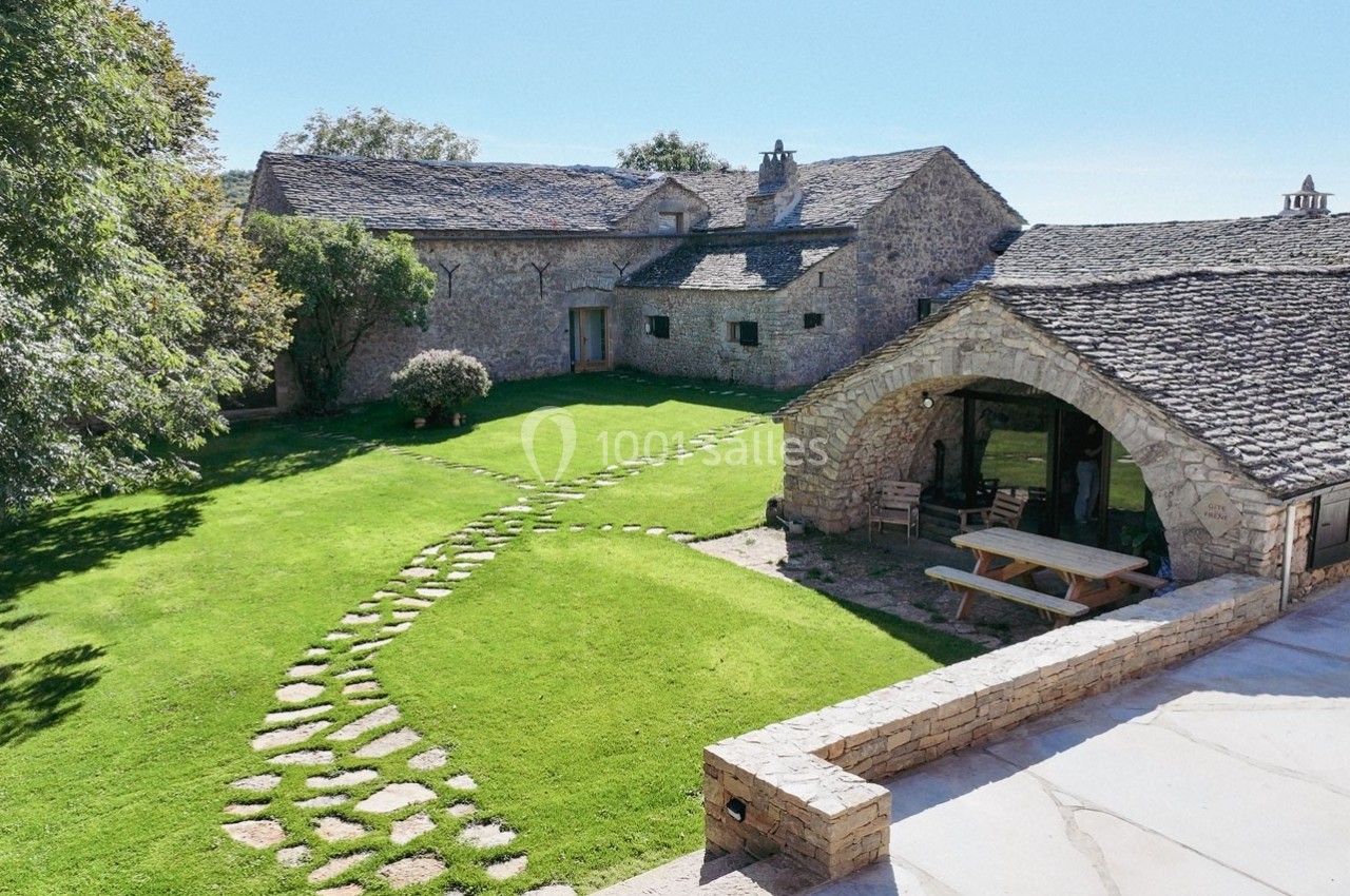 Cour verdoyante avec chemin en pierre, entourée de bâtiments en pierre au toit en ardoise sous un ciel dégagé.