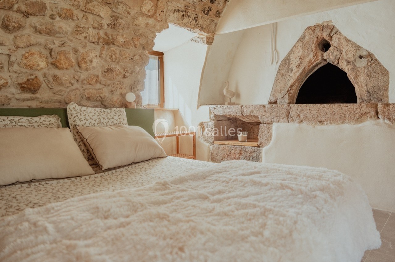 Chambre avec murs en pierre, lit double avec couverture blanche et cheminée ancienne en pierre.