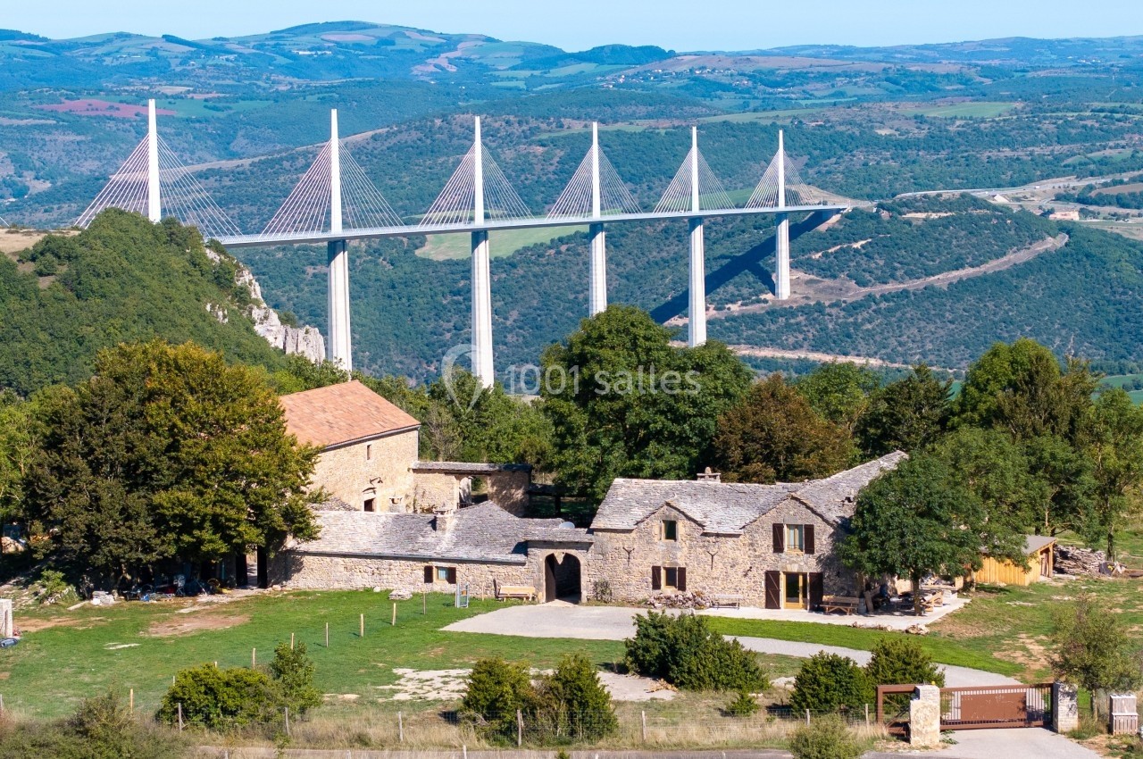 Vue d'un hameau rural avec des bâtiments en pierre, entouré de verdure, et le viaduc de Millau en arrière-plan.