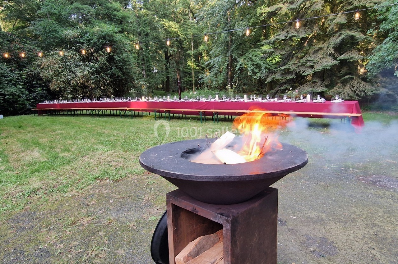 Brasero allumé au premier plan dans un jardin, avec une longue table dressée sous des guirlandes lumineuses en arrière-plan.