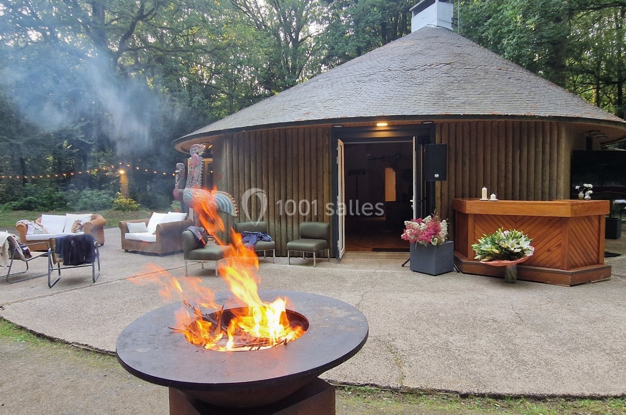 Brasero allumé devant une maison ronde en bois entourée de végétation, avec mobilier de jardin et décoration extérieure.