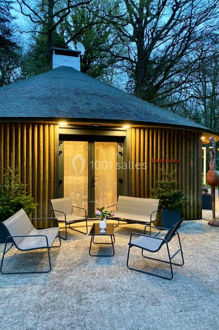 Espace extérieur aménagé avec des fauteuils et une table basse devant une cabane ronde éclairée, entourée d'arbres.