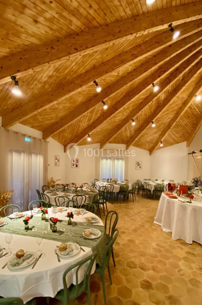 Salle de réception avec tables dressées, chaises vertes, nappes blanches et plafond en bois à poutres apparentes.