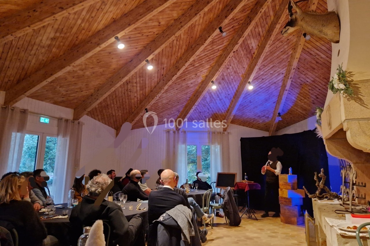Un magicien réalise un tour devant un public assis dans une salle au plafond en bois avec une ambiance chaleureuse.