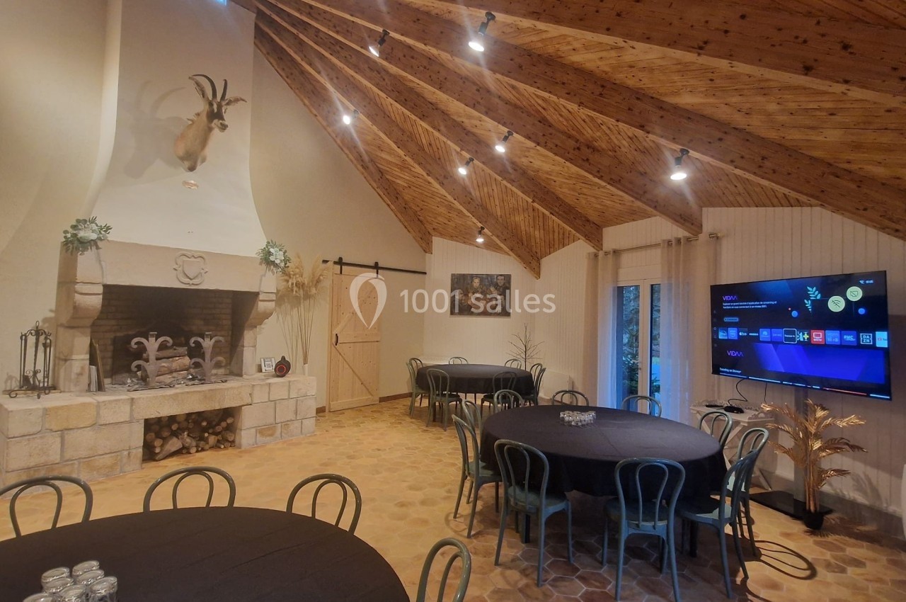 Salle avec tables rondes, cheminée décorative, plafond en bois, et écran mural affichant une interface numérique.