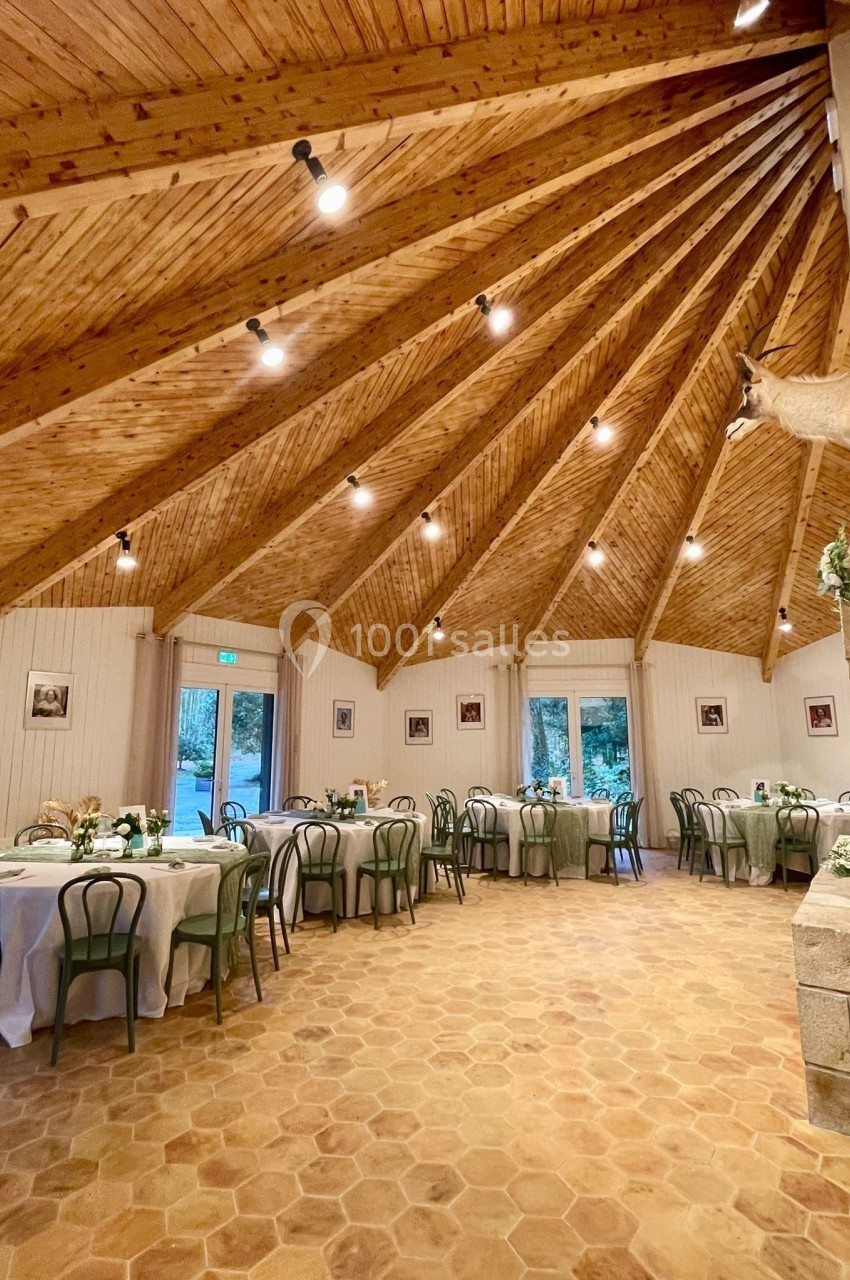 Salle lumineuse avec tables rondes dressées, chaises vertes, plafond en bois en pente et sol en carreaux hexagonaux.