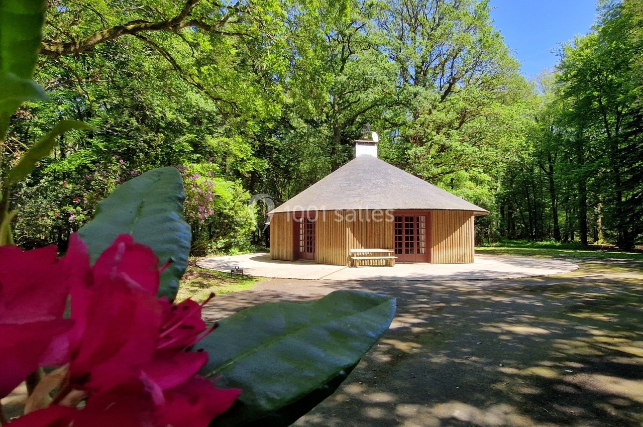Cabane en bois au toit conique entourée d'arbres, avec des fleurs roses au premier plan.
