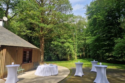 Espace extérieur avec tables hautes dressées et une cabane en bois, entouré d'arbres dans un cadre naturel.
