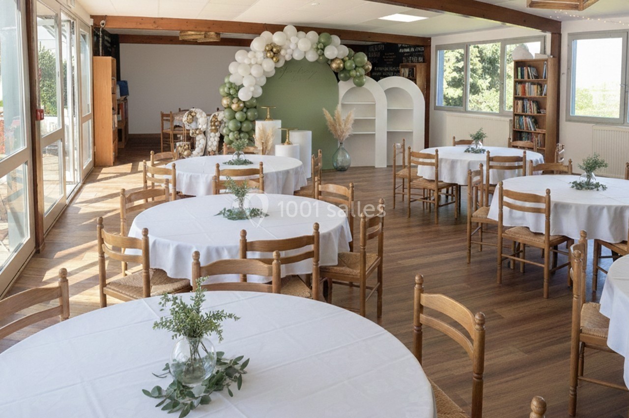 Salle lumineuse avec tables rondes dressées, décorations florales et arche de ballons blancs et verts au fond.