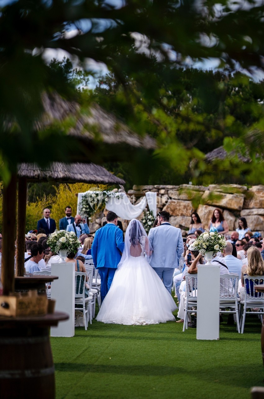 Mariée en robe blanche avançant dans une allée extérieure, entourée d'invités assis, lors d'une cérémonie de mariage.