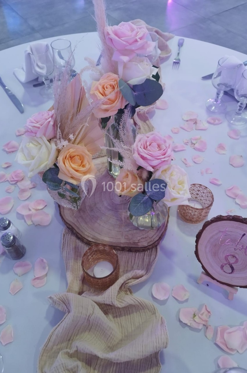 Centre de table avec fleurs pastel, rondins de bois, bougies et pétales roses sur une nappe blanche.