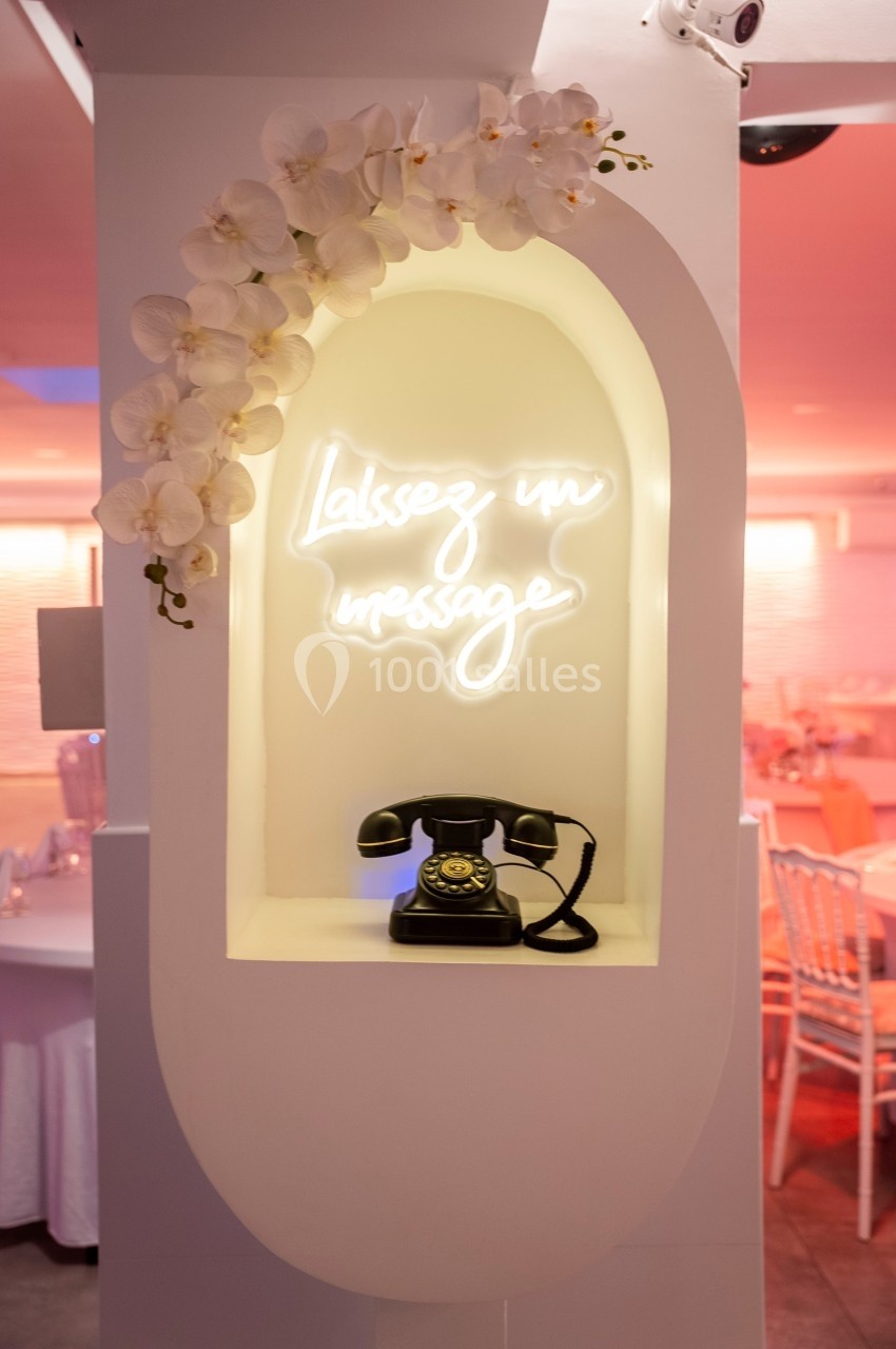 Téléphone vintage noir sous une enseigne lumineuse ’Laissez un message’, encadré par des orchidées blanches.