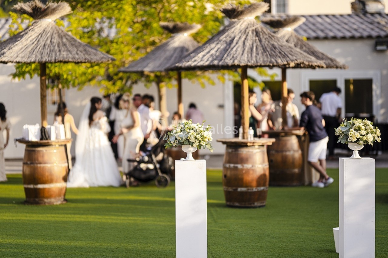 Des compositions florales sur des socles blancs devant une réception en plein air avec des invités et des parasols en paille.