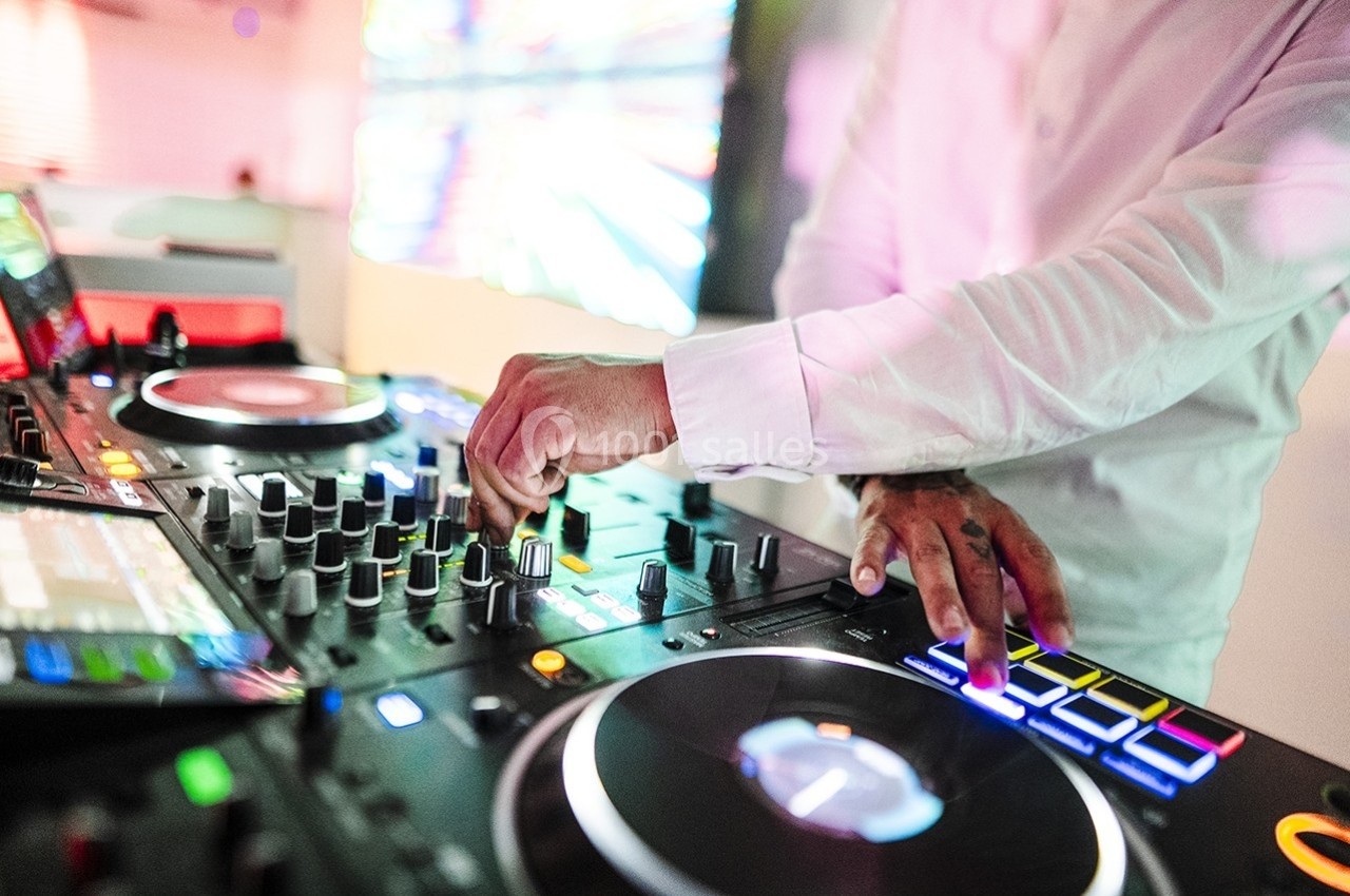 Un DJ en chemise blanche mixe de la musique sur une table de mixage avec des lumières colorées en arrière-plan.