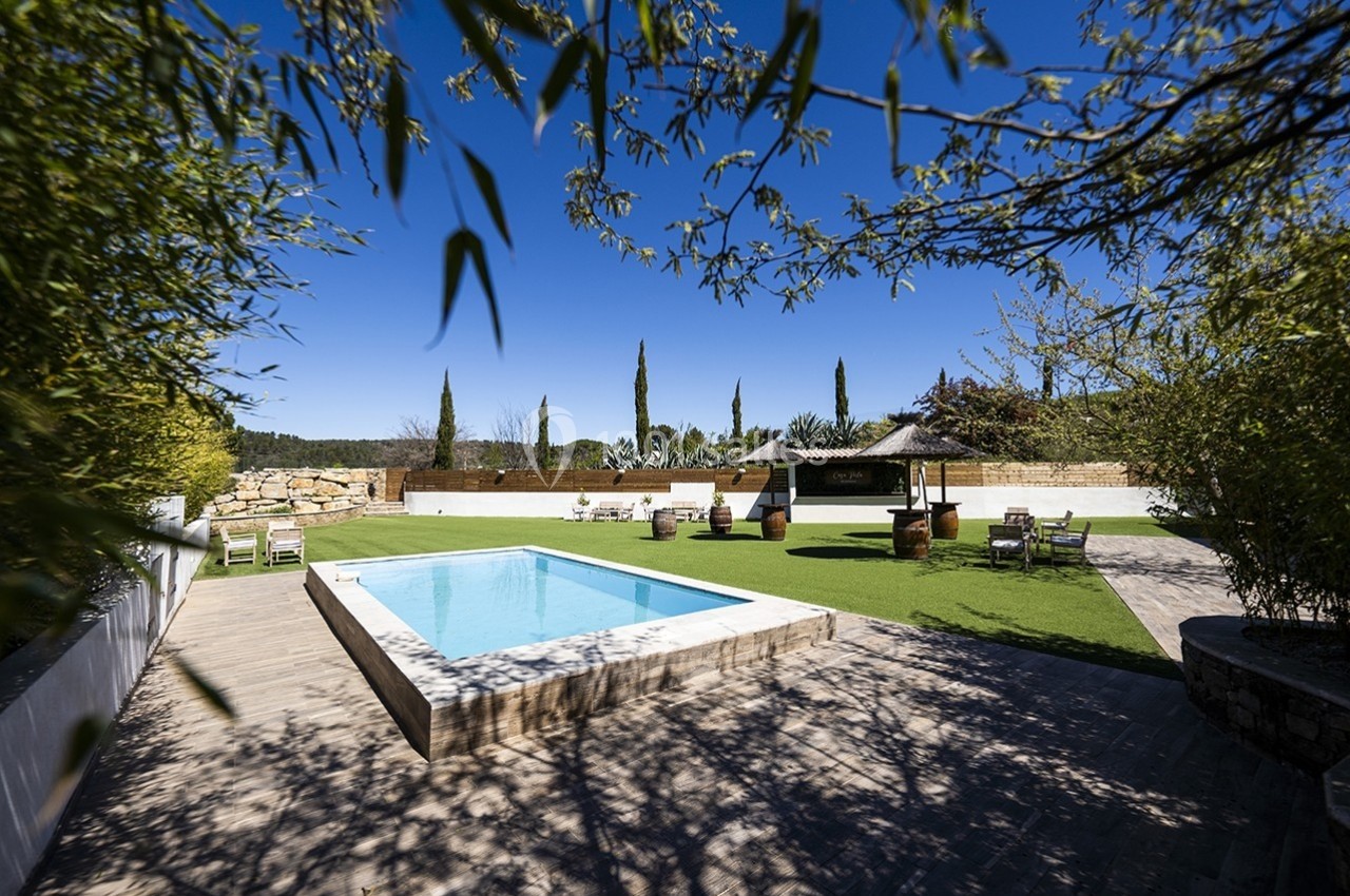 Jardin avec piscine rectangulaire, terrasse en bois, pelouse, mobilier d'extérieur et pergola sous un ciel bleu.