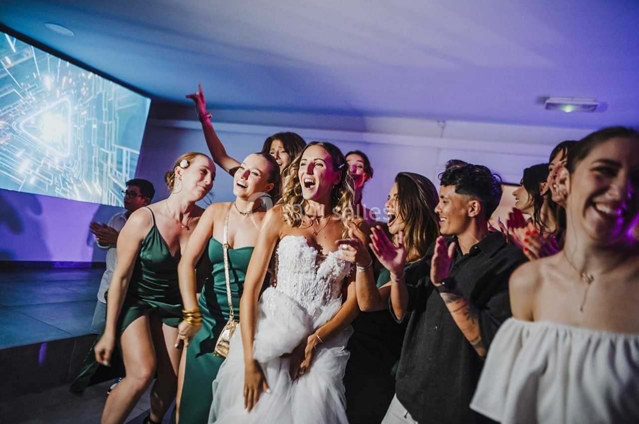 Un groupe de personnes, dont une mariée en robe blanche, dansant et célébrant dans une ambiance festive en intérieur.