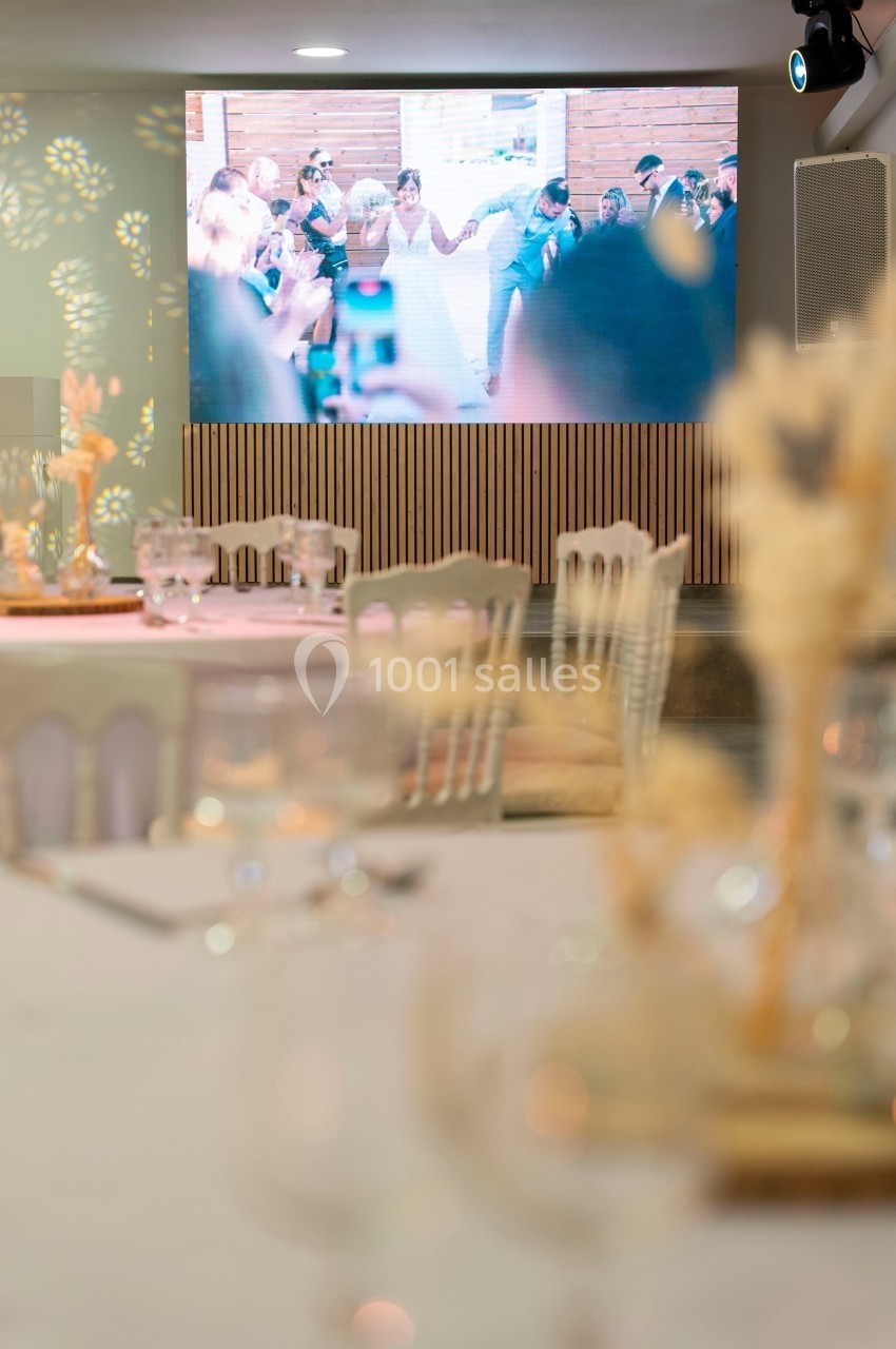 Salle de réception avec tables décorées au premier plan et écran affichant une scène de célébration en arrière-plan.