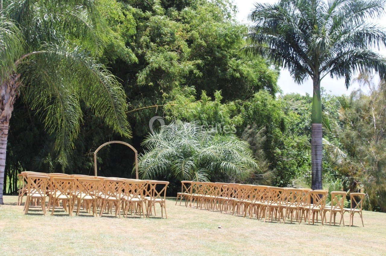 Chaises en bois disposées en rangées sur une pelouse, face à une arche décorative, entourées de palmiers et de verdure.