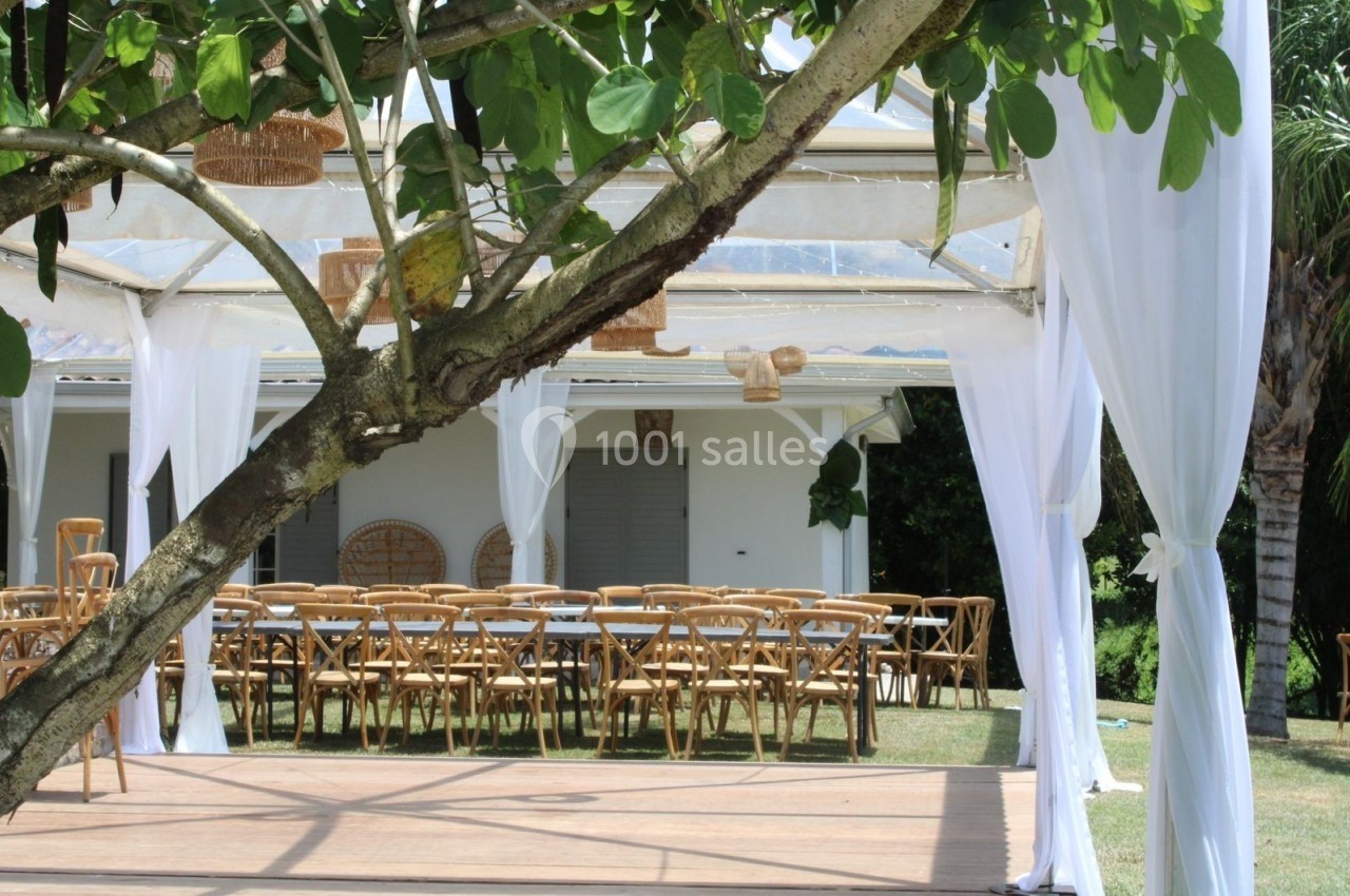 Salle extérieure décorée avec des chaises en bois et des rideaux blancs sous une structure transparente.