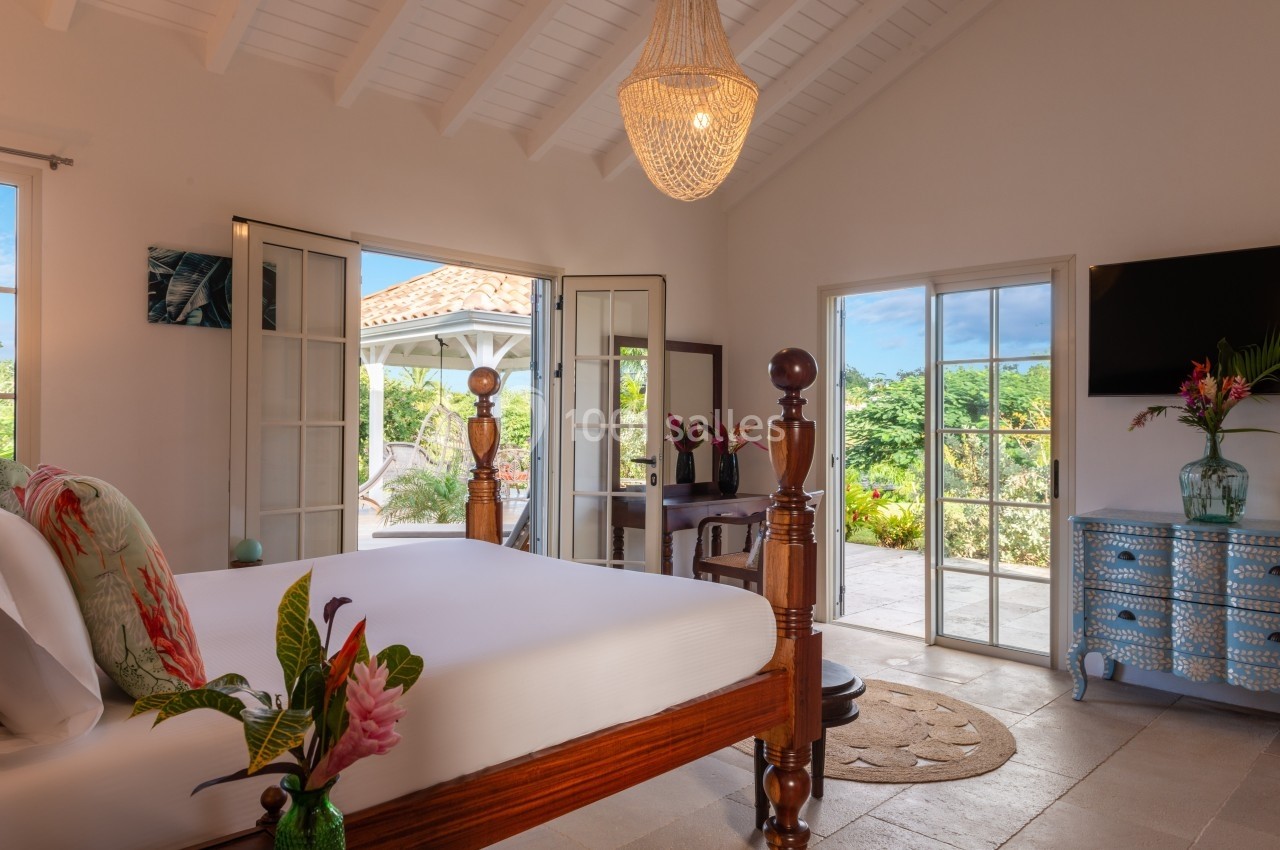 Chambre lumineuse avec lit à baldaquin, décoration tropicale, grandes fenêtres ouvertes sur une terrasse verdoyante.