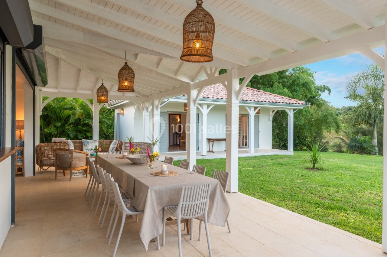 Terrasse couverte avec grande table dressée, chaises blanches, lampes suspendues et vue sur jardin verdoyant.