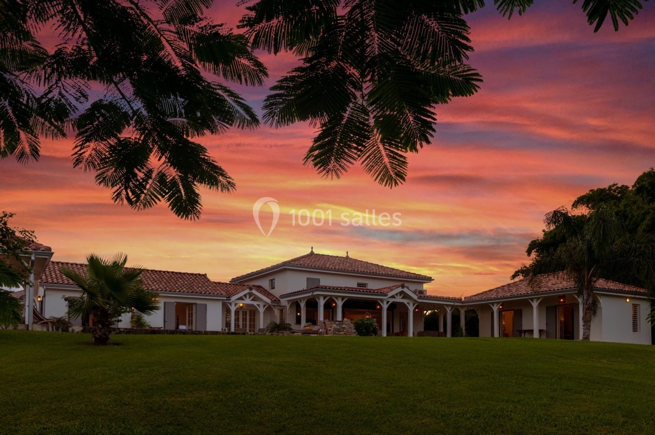 Maison de style méditerranéen avec toit en tuiles, entourée de verdure, sous un ciel coloré au coucher du soleil.