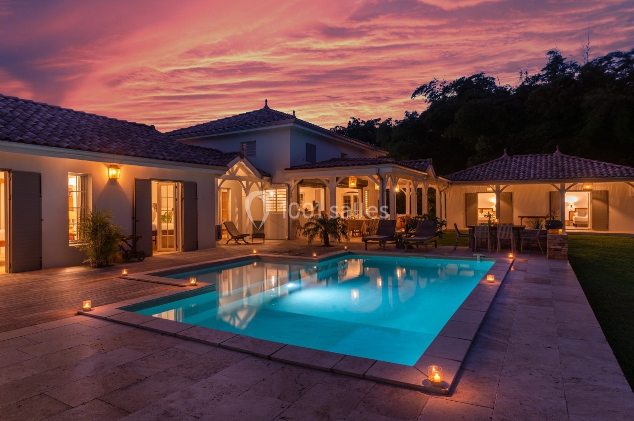 Maison avec piscine éclairée au crépuscule, entourée de terrasses en pierre et de végétation en arrière-plan.