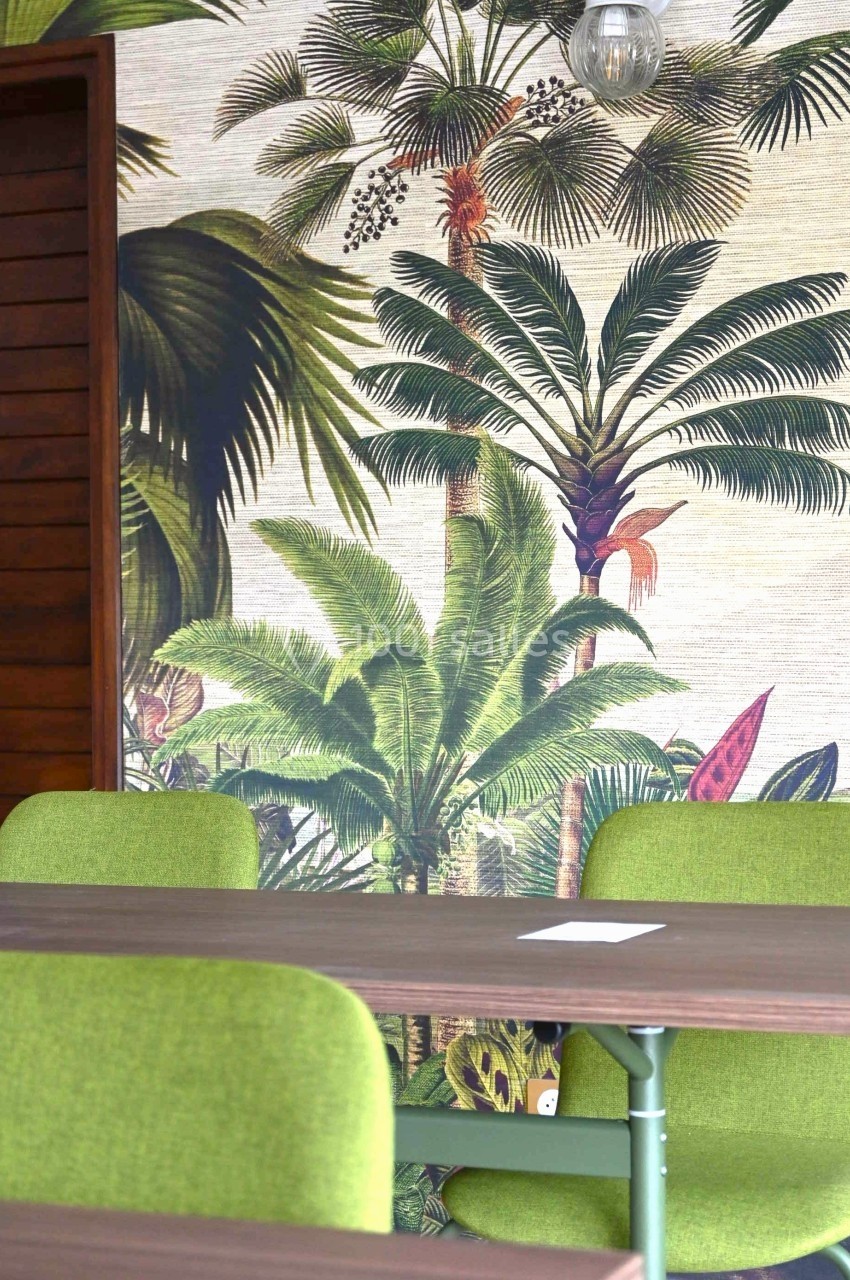 Mur décoré d'un papier peint tropical avec des palmiers, table en bois et chaises vertes au premier plan.